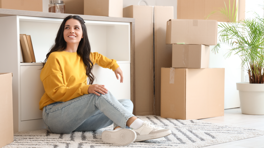 Smiling woman in yellow sweater sitting on floor surrounded by moving boxes in new home