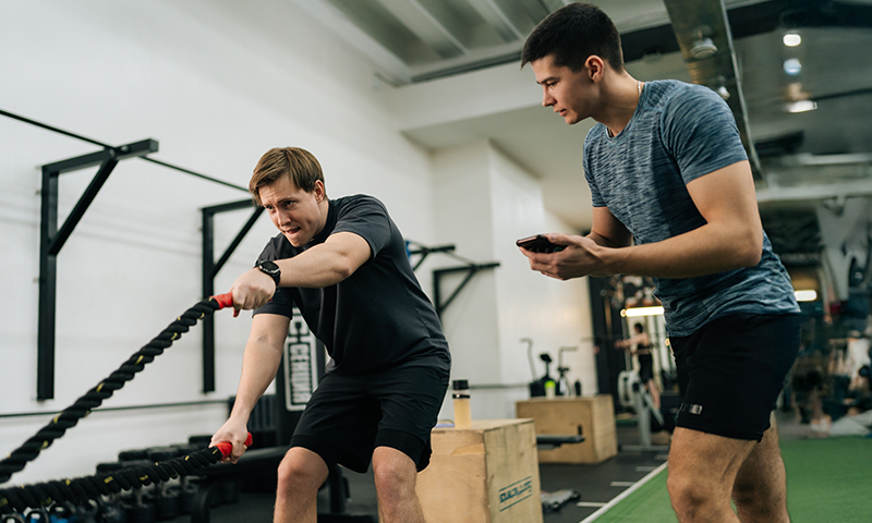 Strength and conditioning coach tracking athlete doing battle rope exercise in gym
