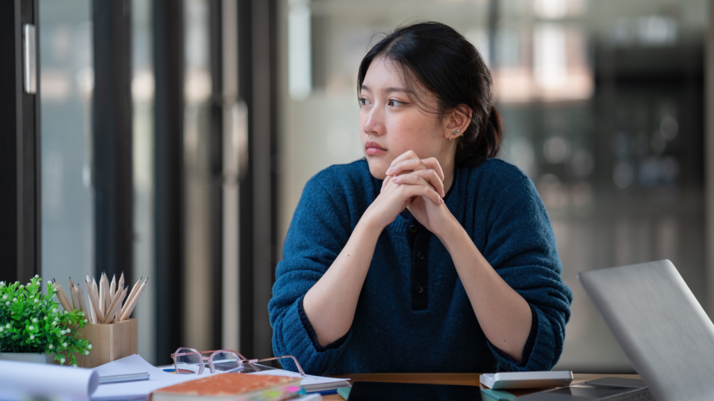 Young Asian woman in blue sweater sitting at office desk with hands clasped, gazing thoughtfully to the side