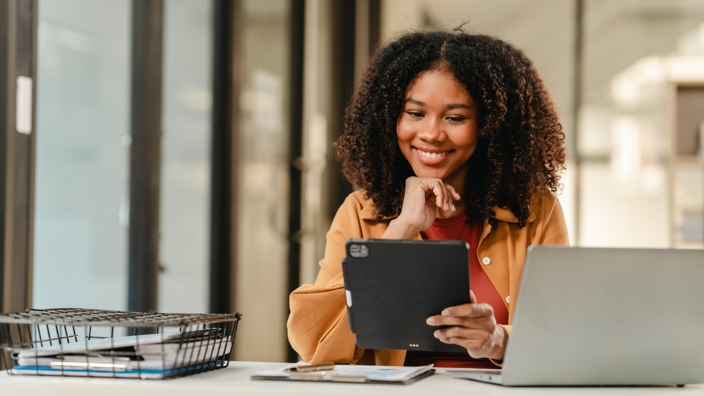 Smiling woman in yellow jacket reviewing tablet at office desk with laptop nearby