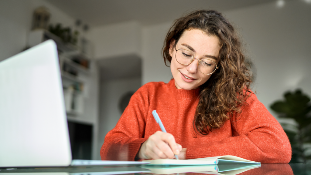 Smiling woman in red sweater writing in notebook beside laptop while studying at home